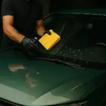 Tecnico applica trattamento idrofobico su parabrezza di auto verde con panno e prodotto specifico